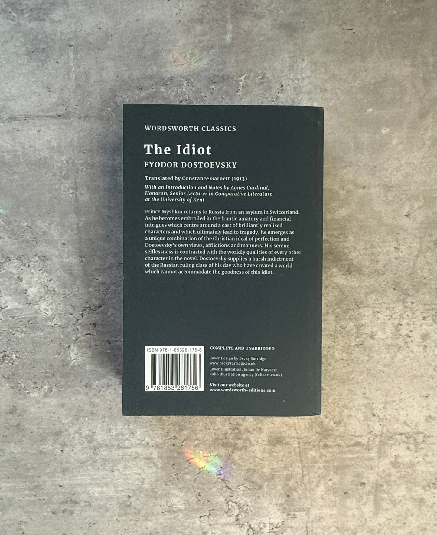 A softcover book of Fyodor Dostoevsky's 'The Idiot' from Wordsworth Classics, featuring a dark cover with a stylized monochrome illustration of a man in a top hat and coat, photographed flat on a textured gray concrete surface. Shop for new and used books with The Stone Circle, the only online bookstore near you in Nevada City, California.