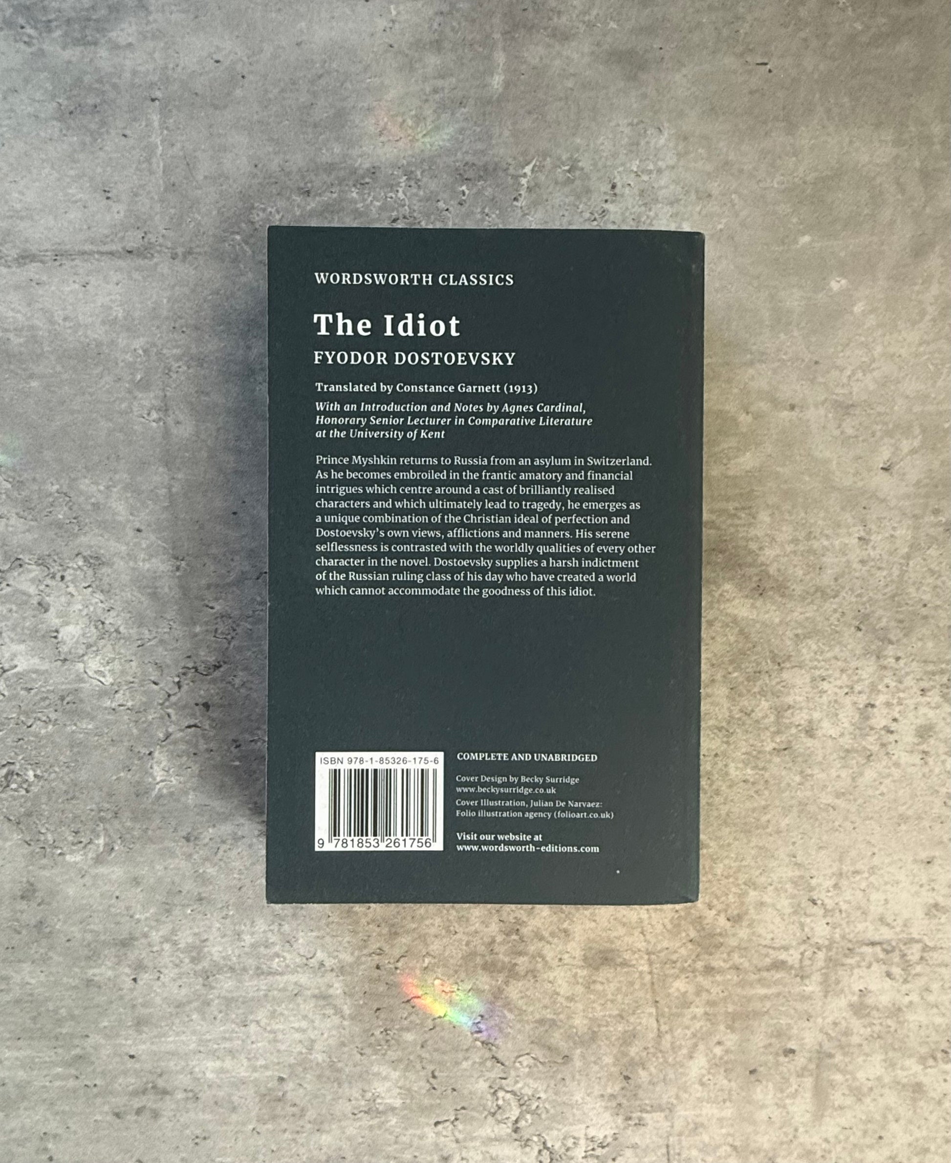 A softcover book of Fyodor Dostoevsky's 'The Idiot' from Wordsworth Classics, featuring a dark cover with a stylized monochrome illustration of a man in a top hat and coat, photographed flat on a textured gray concrete surface. Shop for new and used books with The Stone Circle, the only online bookstore near you in Nevada City, California.