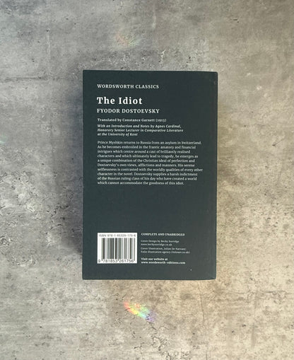 A softcover book of Fyodor Dostoevsky's 'The Idiot' from Wordsworth Classics, featuring a dark cover with a stylized monochrome illustration of a man in a top hat and coat, photographed flat on a textured gray concrete surface. Shop for new and used books with The Stone Circle, the only online bookstore near you in Nevada City, California.