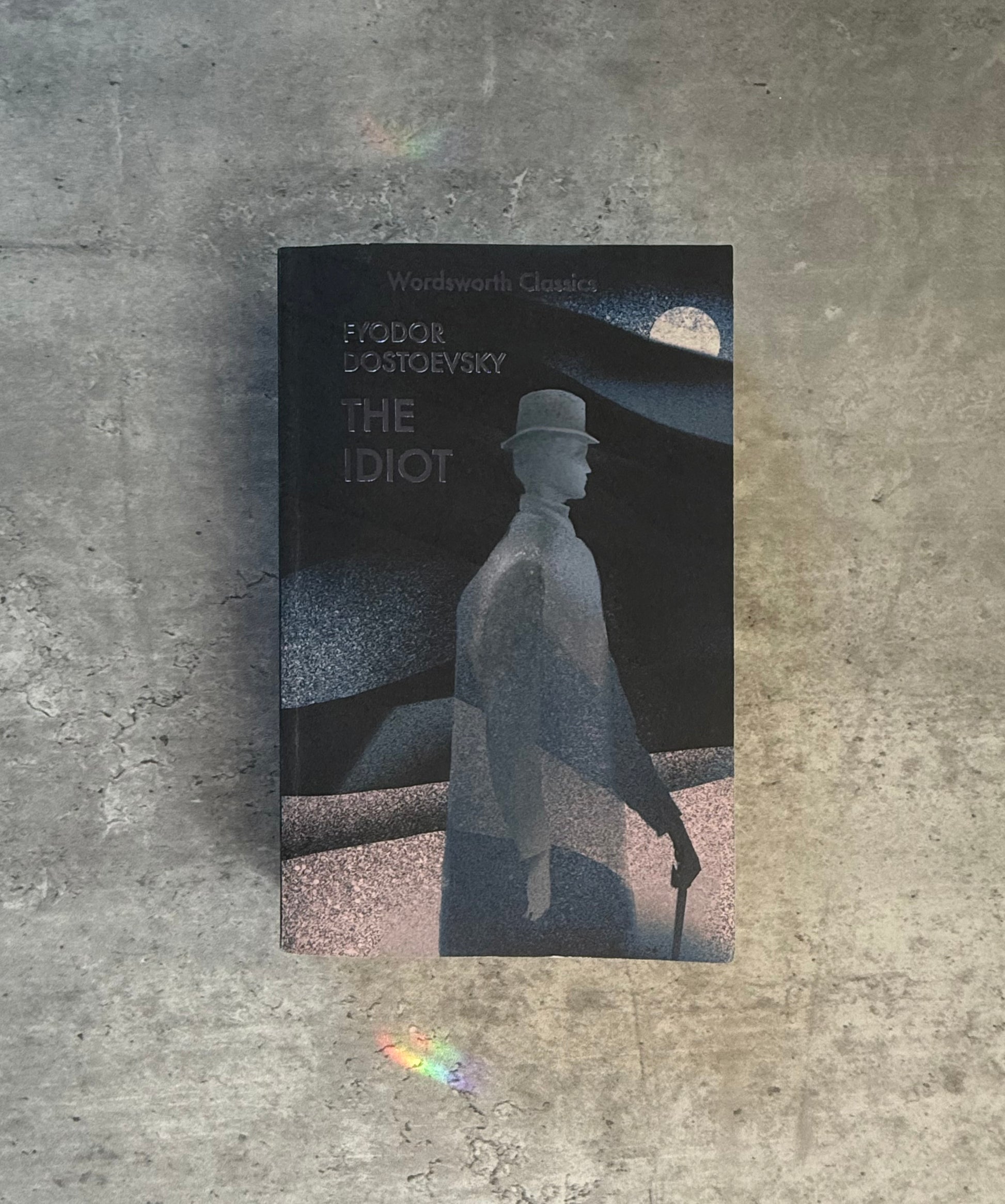 A softcover book of Fyodor Dostoevsky's 'The Idiot' from Wordsworth Classics, featuring a dark cover with a stylized monochrome illustration of a man in a top hat and coat, photographed flat on a textured gray concrete surface. Shop for new and used books with The Stone Circle, the only online bookstore near you in Nevada City, California.