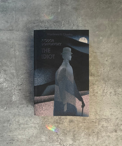 A softcover book of Fyodor Dostoevsky's 'The Idiot' from Wordsworth Classics, featuring a dark cover with a stylized monochrome illustration of a man in a top hat and coat, photographed flat on a textured gray concrete surface. Shop for new and used books with The Stone Circle, the only online bookstore near you in Nevada City, California.