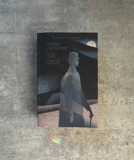 A softcover book of Fyodor Dostoevsky's 'The Idiot' from Wordsworth Classics, featuring a dark cover with a stylized monochrome illustration of a man in a top hat and coat, photographed flat on a textured gray concrete surface. Shop for new and used books with The Stone Circle, the only online bookstore near you in Nevada City, California.