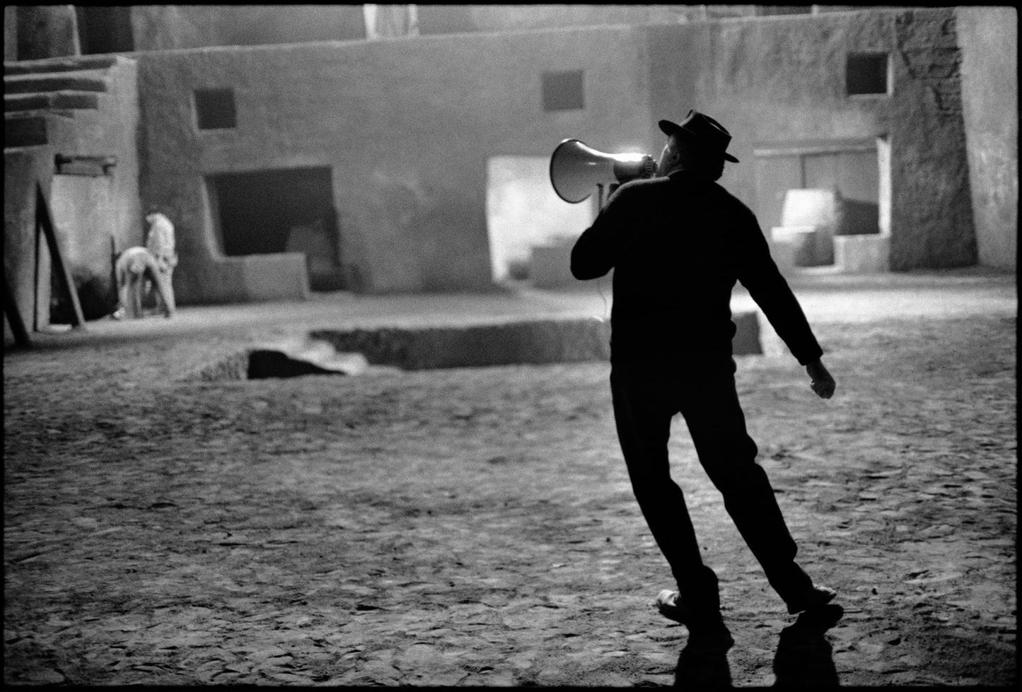 Black and white photograph of Fellini on the set of Fellini Satyricon holding a megaphone in a dusty courtyard, with low-rise buildings in the background.